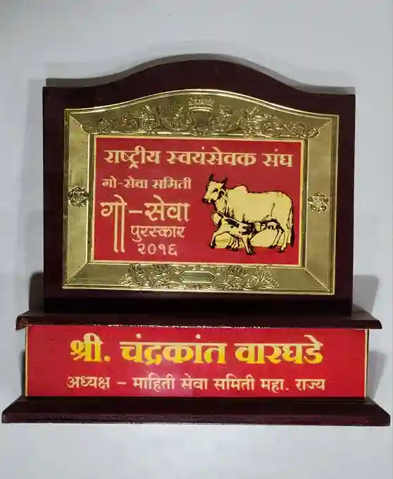 At the center, there is a golden plate with intricate designs, displaying the (Rashtriya Swayamsevak Sangh) and (Go-Seva Puraskar 2022), indicating it is an award for service related to cow welfare.
The plaque also includes an illustration of a cow and calf, symbolizing the theme of the award. Below the central plate, there is a red nameplate that reads (Shri. Chandrakant Waghade), identifying the recipient, along with the title (President - Women's Service Committee, Maharashtra).