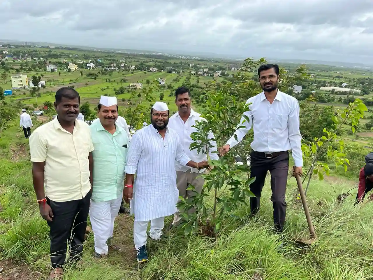 a group of five individuals standing together on a hillside, participating in a tree planting event. They are smiling and posing near a young tree that has been planted.